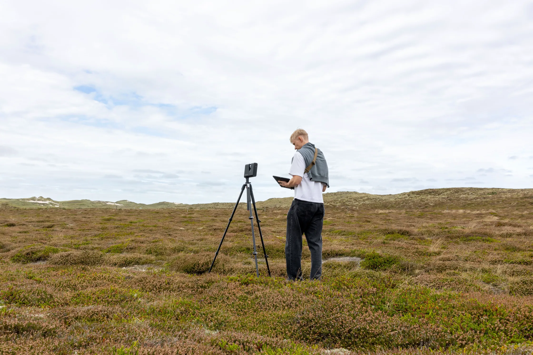 En person sidder på en stol foran et skrivebord med en computer, mens solen skinner ind gennem et vindue.